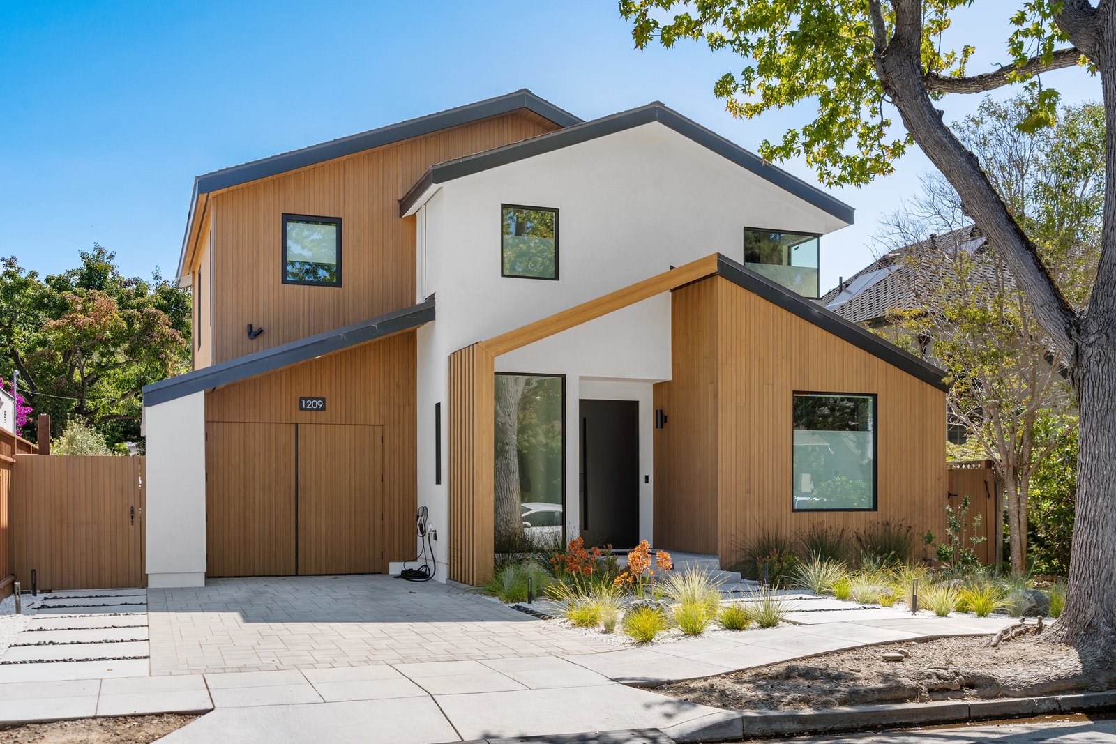 Cabrillo Avenue front exterior: a two-volume modern home with cedar siding and white stucco, peaked metal-clad roof, native planting along the entry walk.