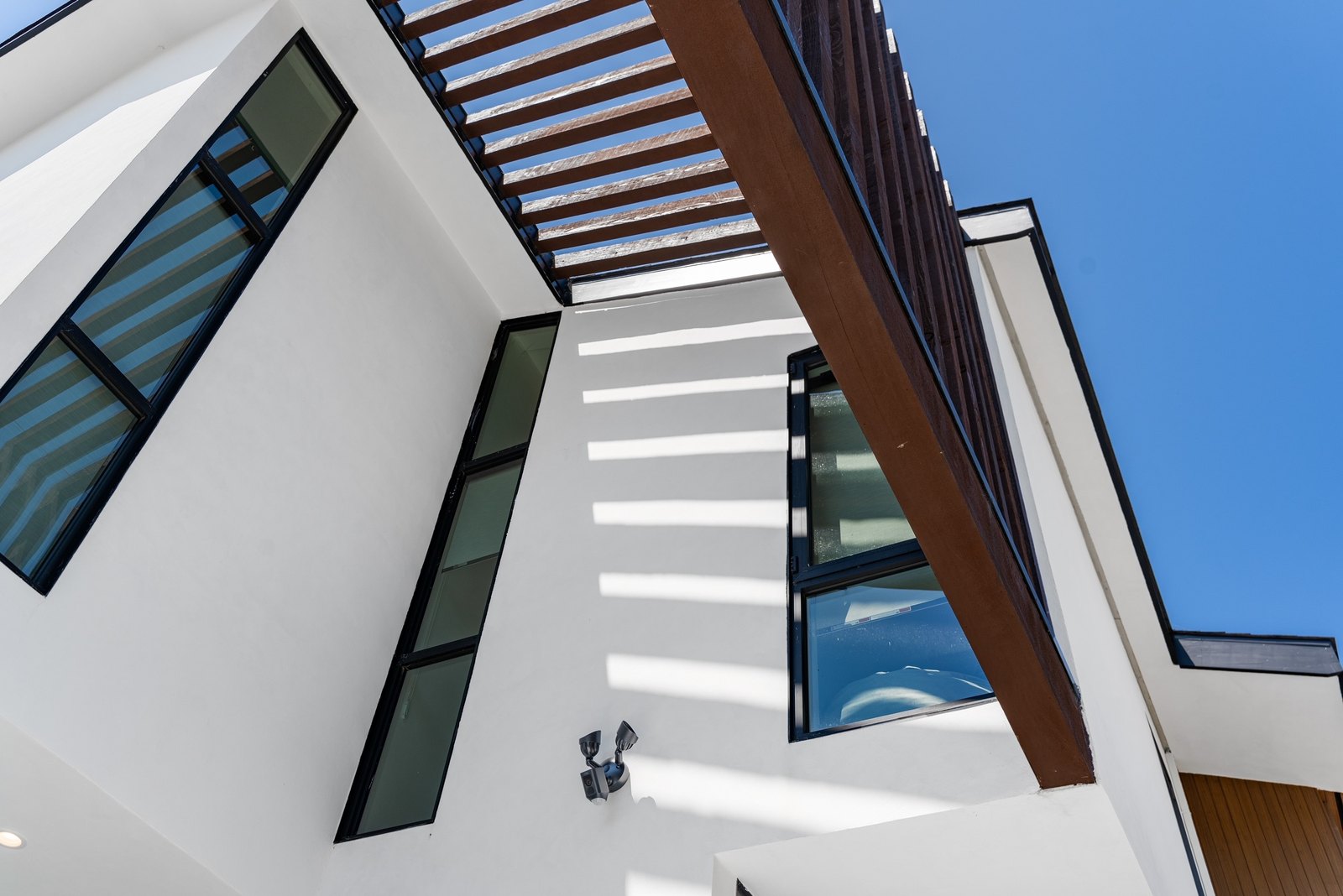 Looking up at a slatted wood pergola against a clear blue sky, set between two angled stucco volumes. Sunlight stripes the white wall below.