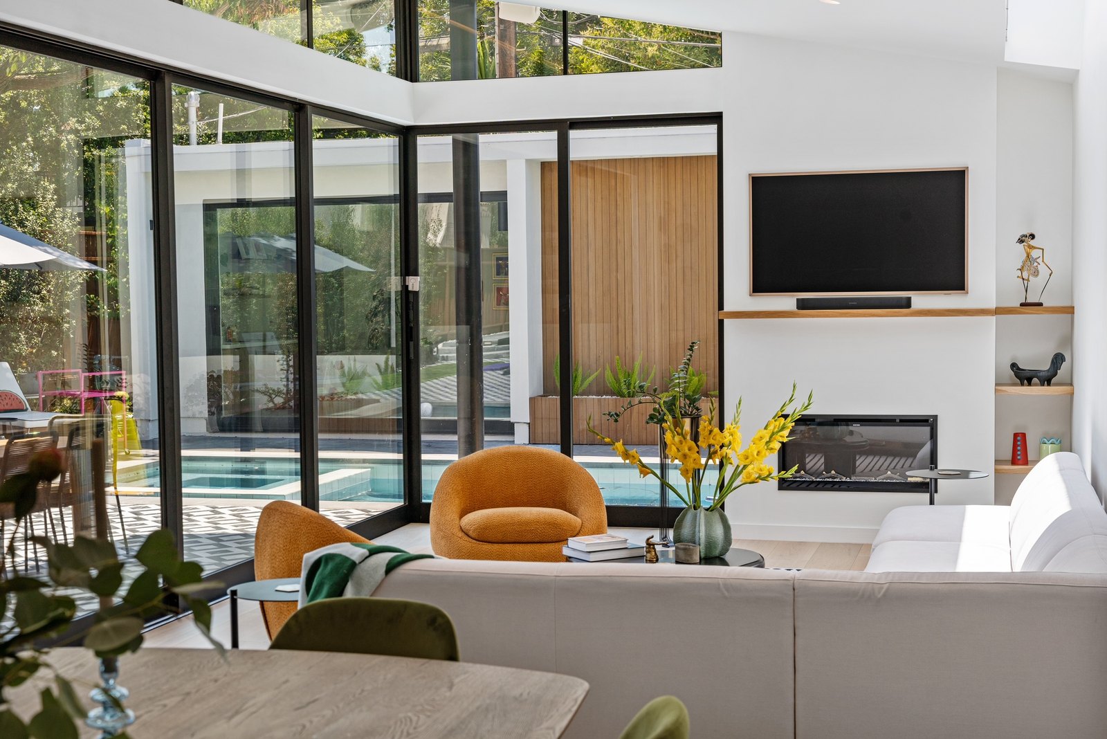 Living room looking out through floor-to-ceiling glazing to the pool: cream sectional, orange swivel chairs, fireplace and inset TV on the far wall.