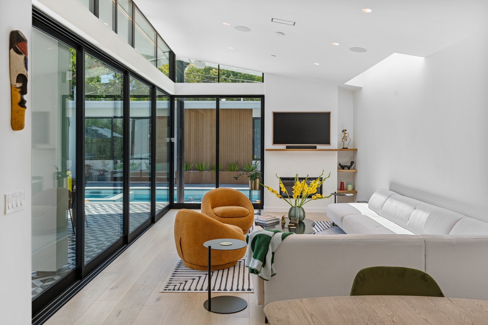 Living room from the dining table side: tall glazing to the pool at left, fireplace and TV ahead, daylight from a clerestory at right.