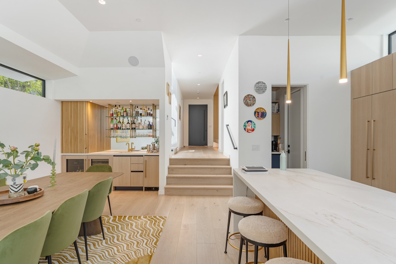Dining and kitchen looking down a corridor: oak table with green velvet chairs at left, wet bar with slatted oak cabinetry, marble counter and brass pendants at right.