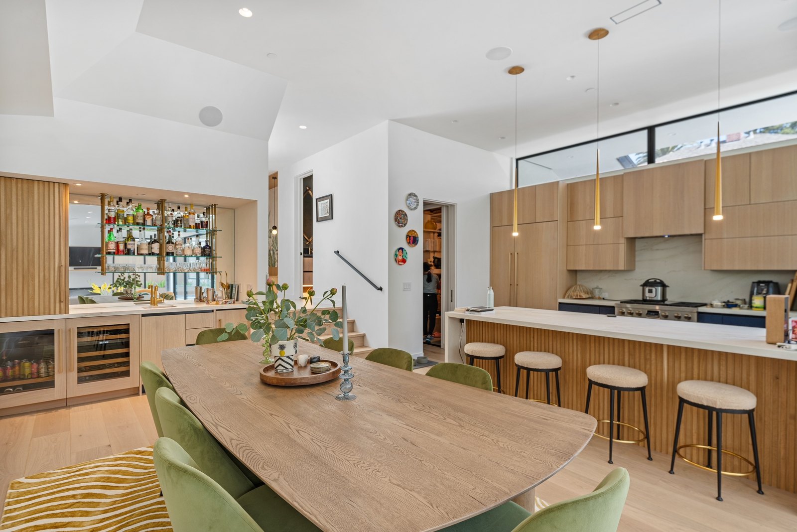 Same room turned to the kitchen: dining table forward, slatted oak island with bar stools, marble counter, induction range, brass pendants.