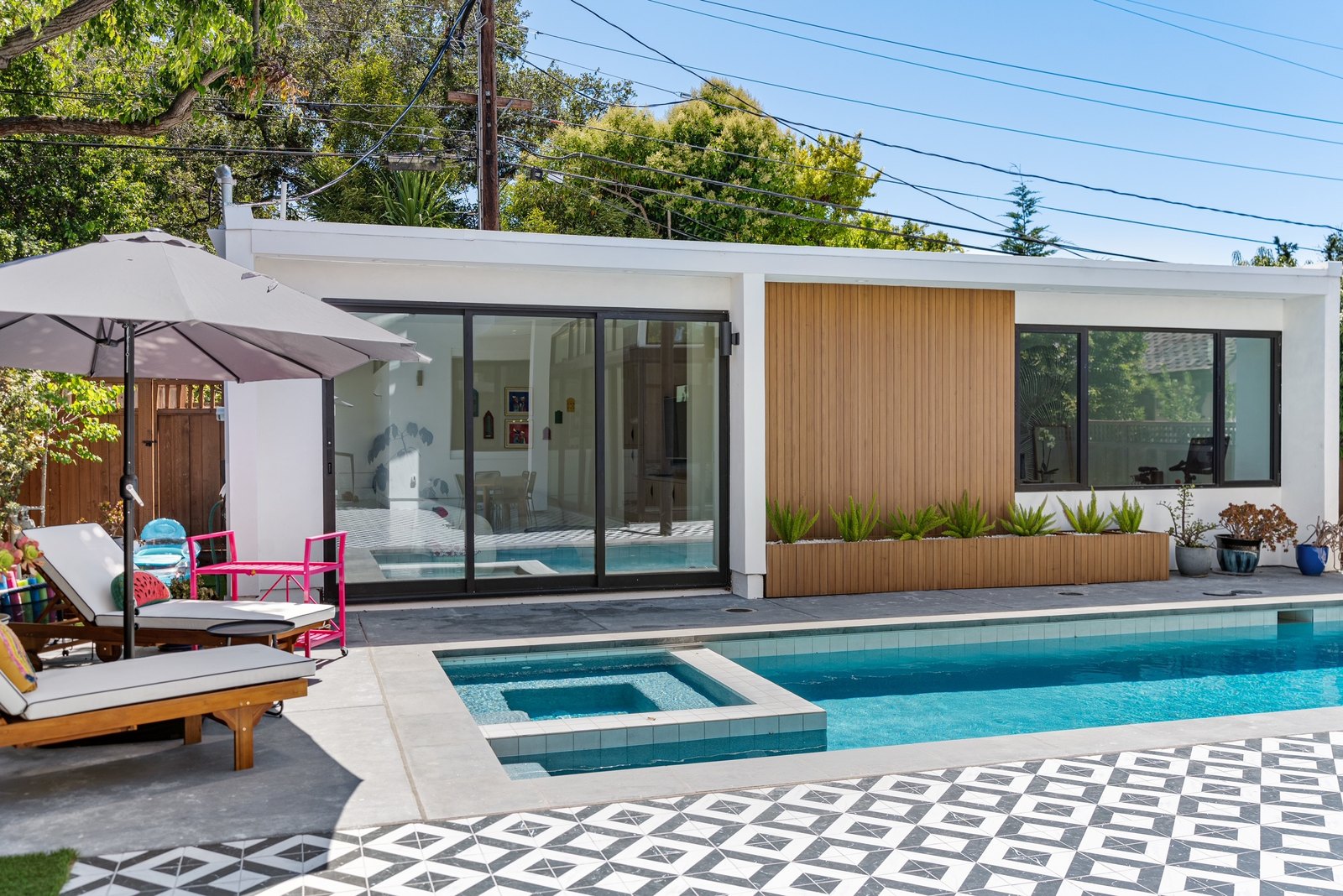 Backyard with pool and spa from the rear: the white-stucco detached ADU with cedar slat panels, geometric checker-tile deck, raised planters along the back wall, umbrella and pink chairs to one side.