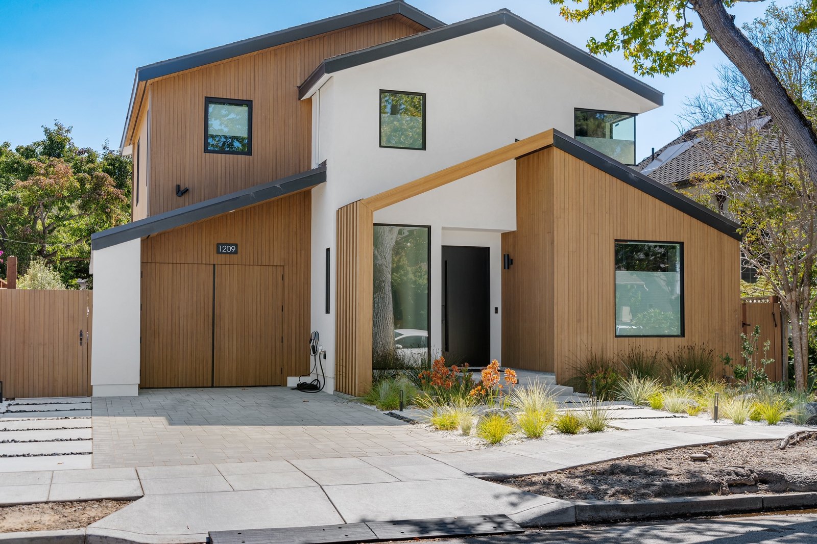 Front exterior at street level: the two-volume modern home with cedar siding and white stucco, peaked roof against sky, entry path with native grasses.