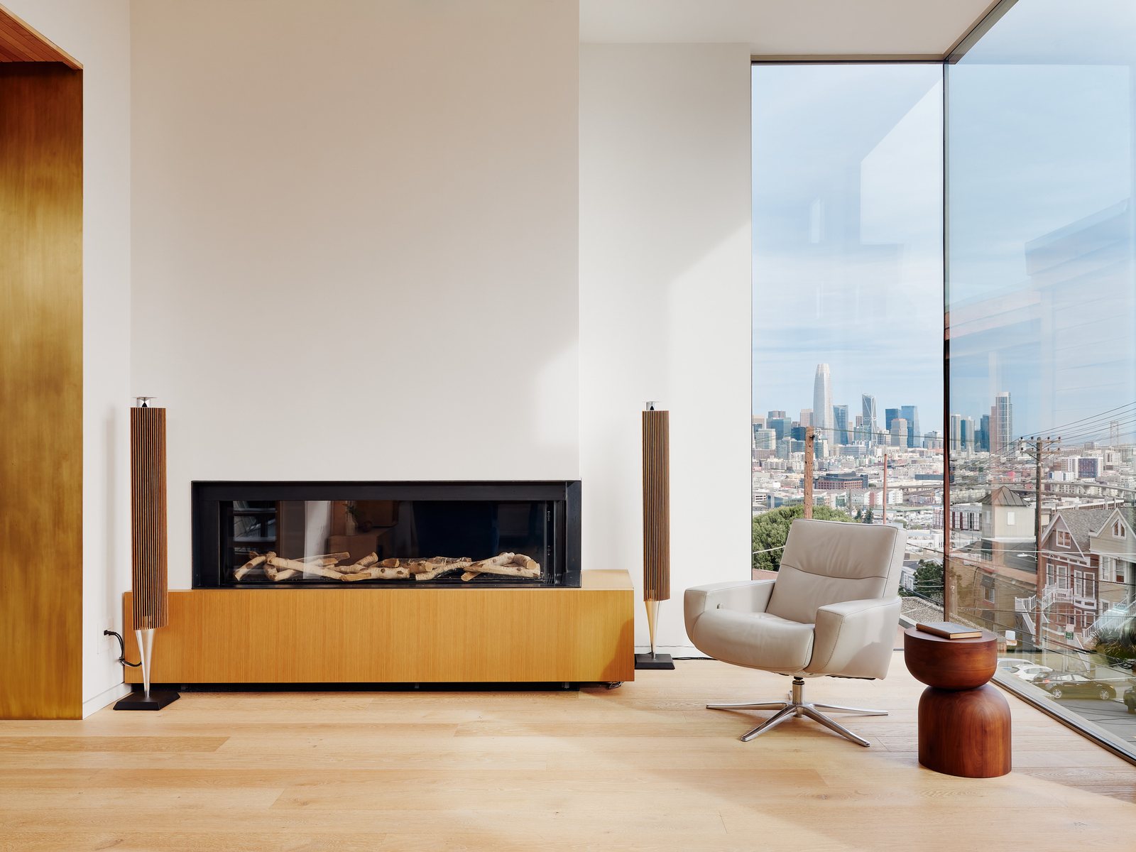 Living room with a linear Ortal fireplace, a swivel chair, BeoLab speakers, and floor-to-ceiling glazing framing the San Francisco skyline.