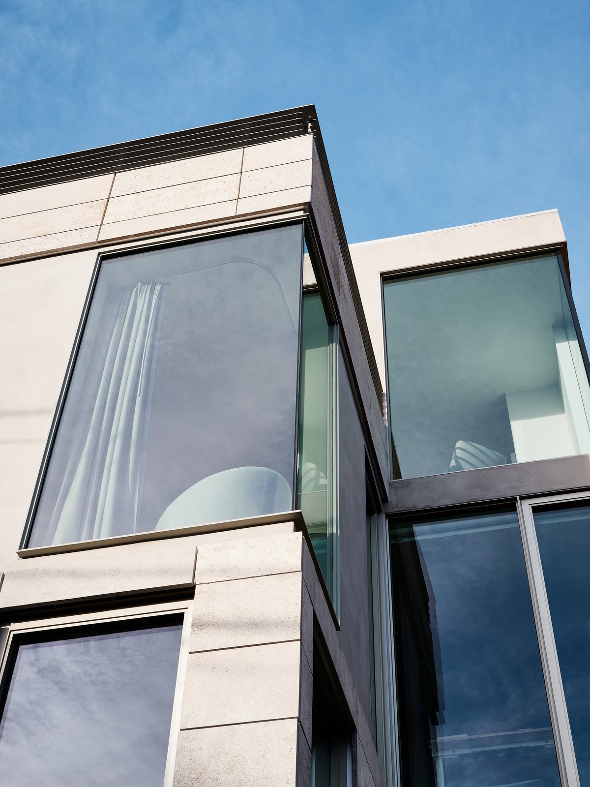 Upper-floor exterior detail: stone rainscreen panels meeting a black-framed mitered corner window against blue sky.