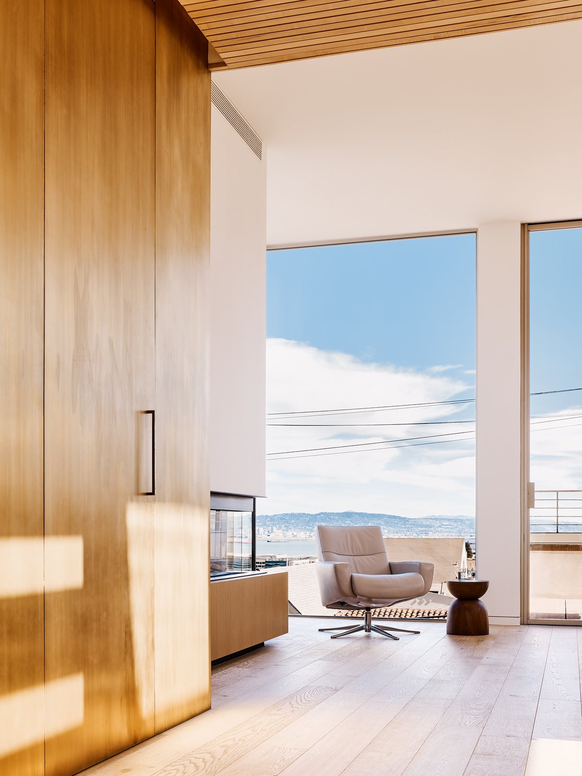 Sitting nook in a wood-paneled corner: single armchair beside floor-to-ceiling glass opening onto the San Francisco hills in afternoon light.