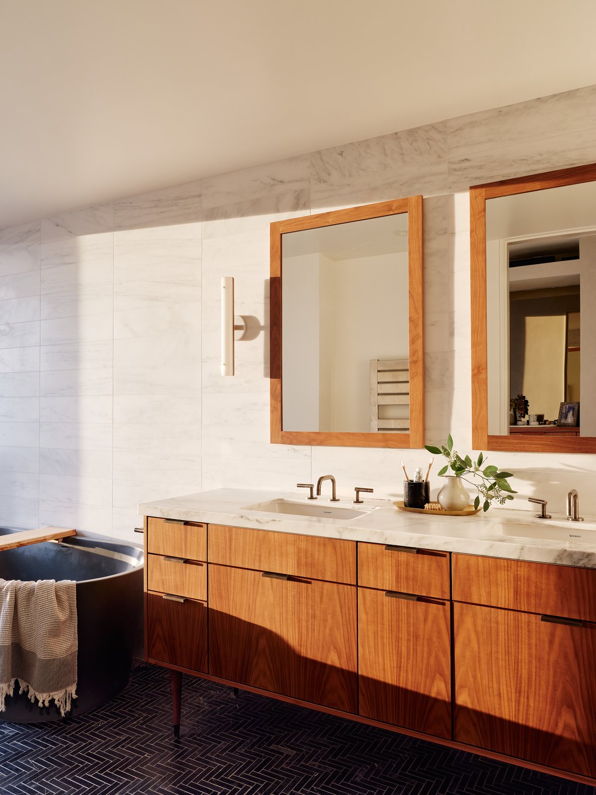 Primary bath with oak vanity, marble counter and backsplash, twin framed mirrors, and a copper free-standing soaking tub at left.