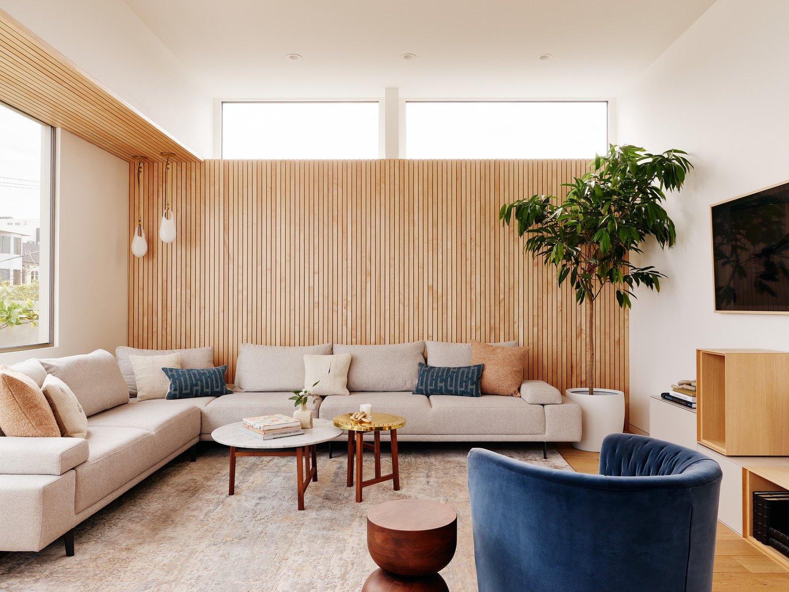 Secondary lounge: a long cream sectional against a floor-to-ceiling wood-slat wall, a blue velvet swivel chair in the foreground, a fiddle-leaf fig at right.
