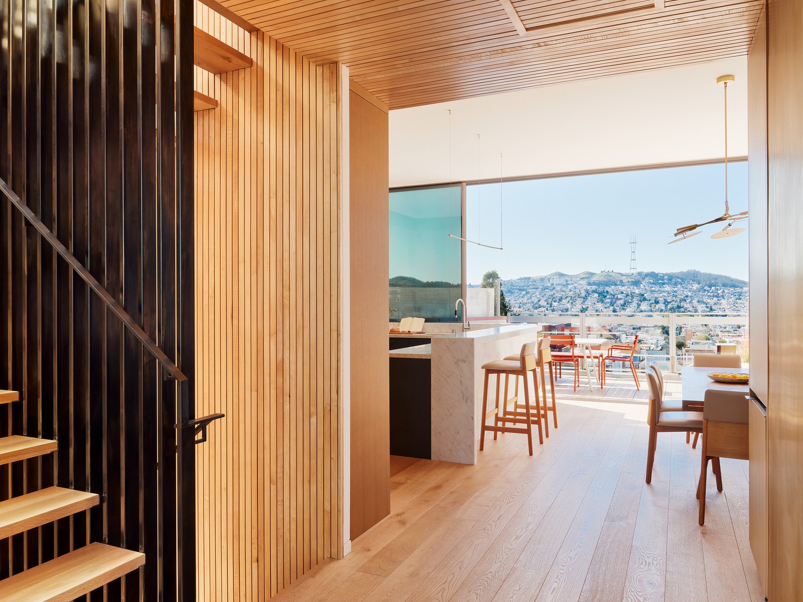 Standing at the top of the open-stair: cedar slat enclosure to the left, the kitchen's marble counter and bar stools at center, the dining table beyond, Twin Peaks visible through the terrace doors.