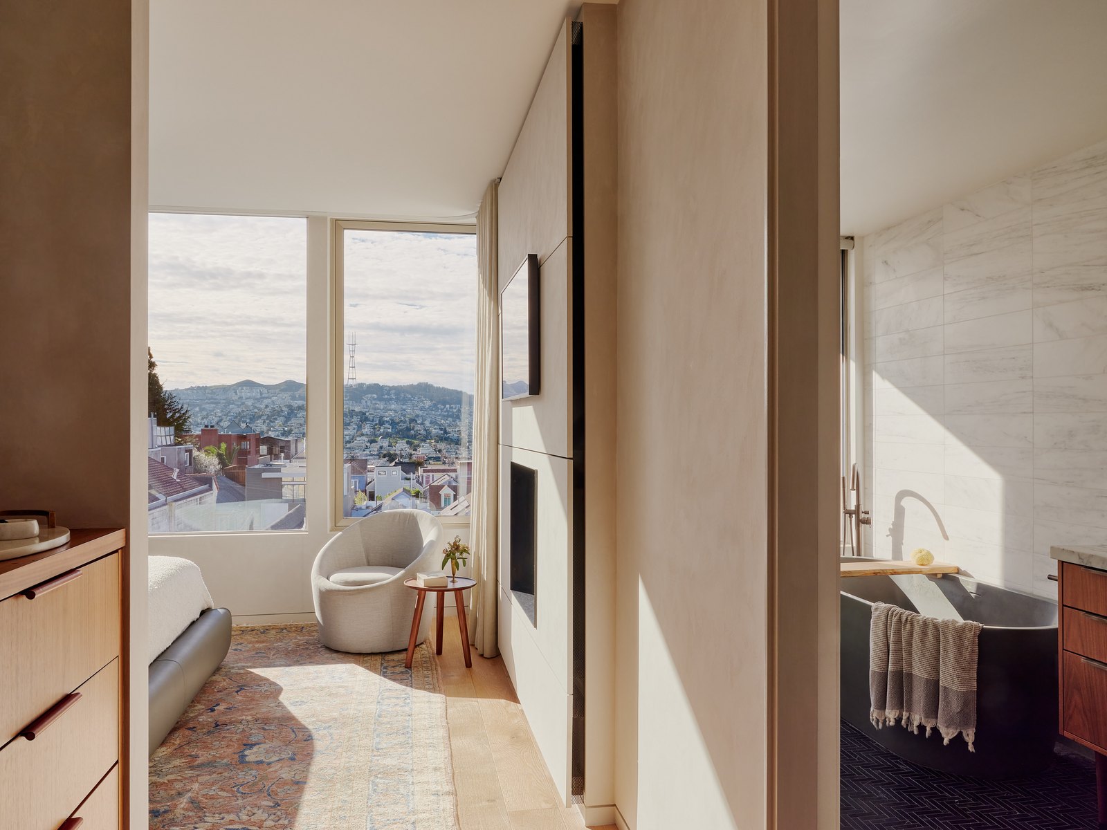 Primary suite seen through an interior aperture: a swivel chair beside a corner window holding the San Francisco hills, the bathroom partly visible at right with a copper soaking tub.