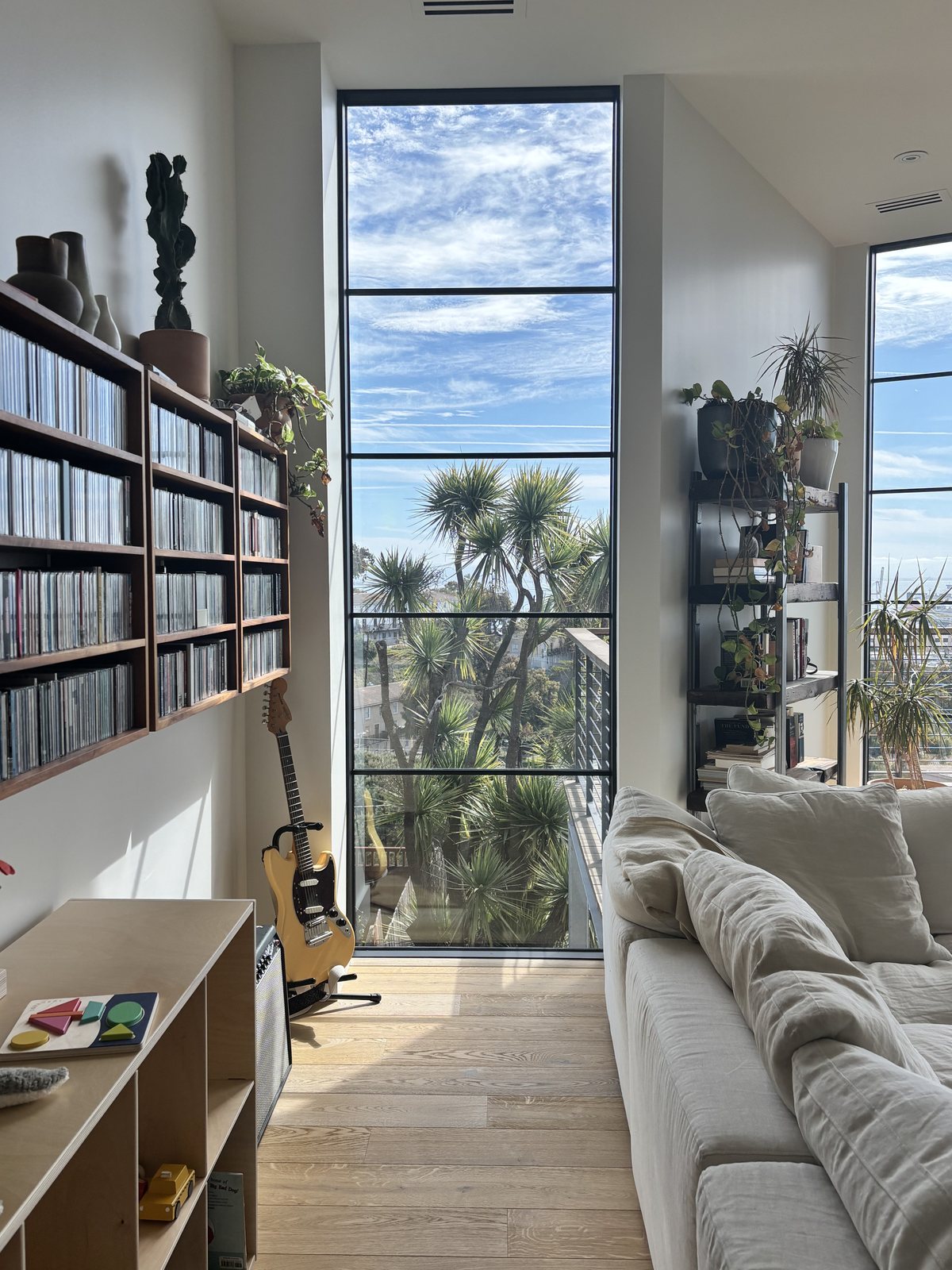 Living room with steel-framed glazing, a long sofa, a wall of vinyl records, an electric guitar, and views to a planted balcony.