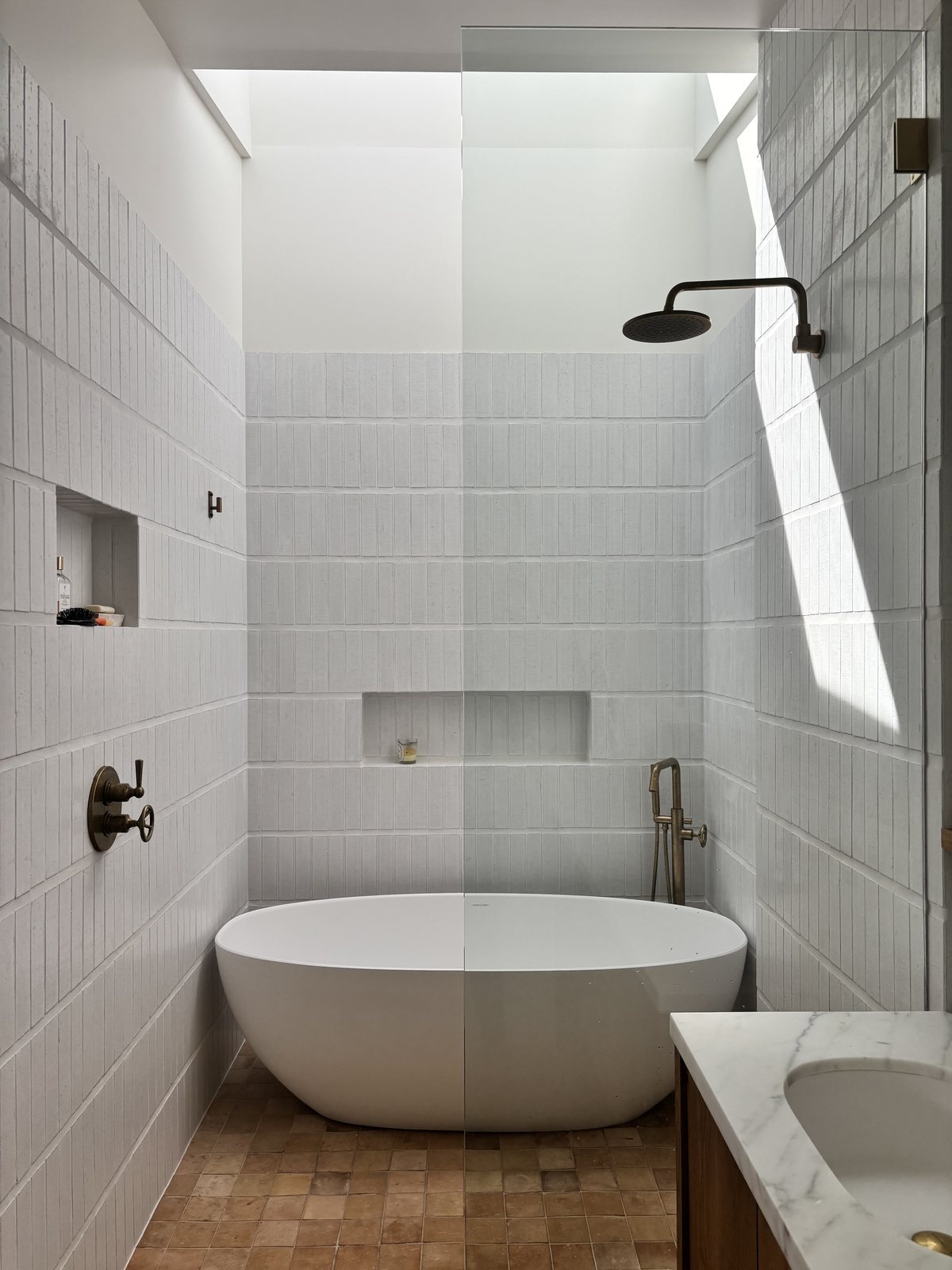 Primary bathroom in minimal white square tile: walk-in shower with bronze fixtures, freestanding white tub, a linear skylight crossing the upper-left corner.