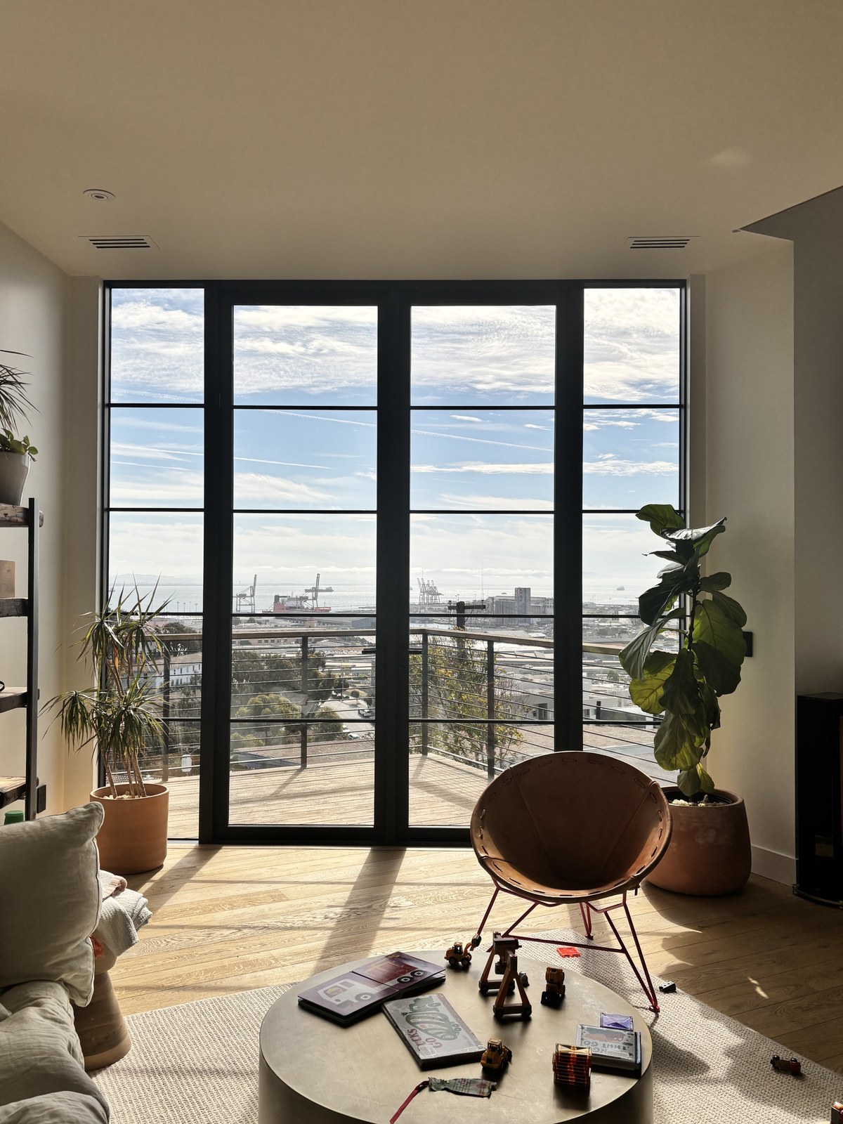 Living room looking out: full-height steel-framed glazing, a fiddle-leaf fig in a terracotta pot, a copper wire chair, and views across the San Francisco Bay to the port cranes.